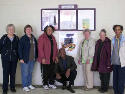 Six women stand to the right and left of a squatting man in front of framed art on a wall. 